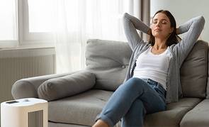 Mujer descansa en un sillón gris con un purificador de aire funcionando a sus pies.