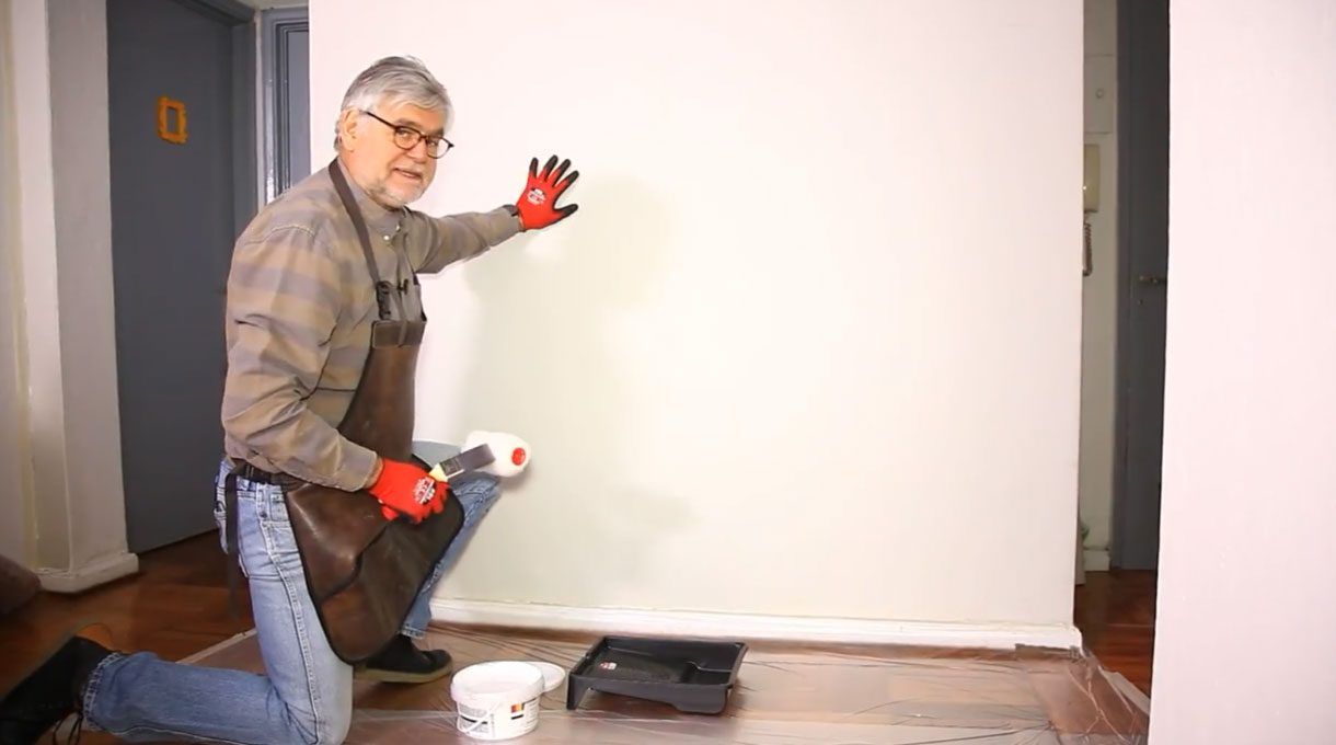 Tardel arrodillado en el piso, sosteniendo un rodillo para pintar en una mano y la otra apoyada en un muro blanco