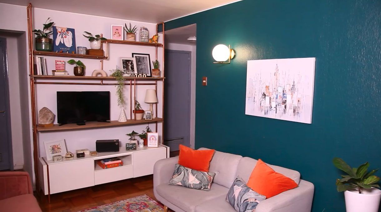 living con el estante de cobre terminado, con un televisor en una repisa y en las otras, objetos decorativos. También hay un sillón de dos cuerpos con cojines naranjos, y un cuadro en la pared verde que fue pintada para el proyecto. Al lado del curadro, un apliqué encendido.
