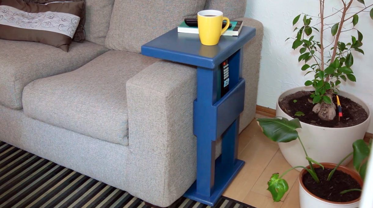 mesa de arrimo instalada en el brazo de la sofá, con libro, control remoto en e bolsillo y una taza de café sobre la mesa
