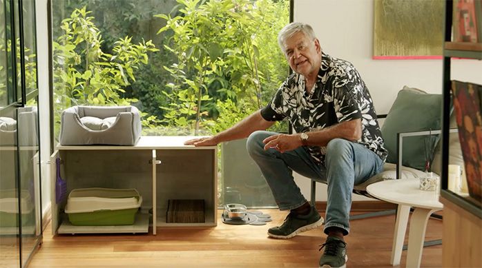 Una persona muestra el mueble arenero para gatos con un arenero y rascador en su interior, una cama para mascotas en la parte superior y un tapete para mascotas con comedero y bebedero para mascota