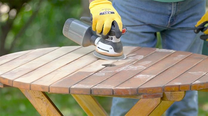 una persona con guantes lija la mesa de madera con una lijadora roto orbital para hacer una restauración de muebles