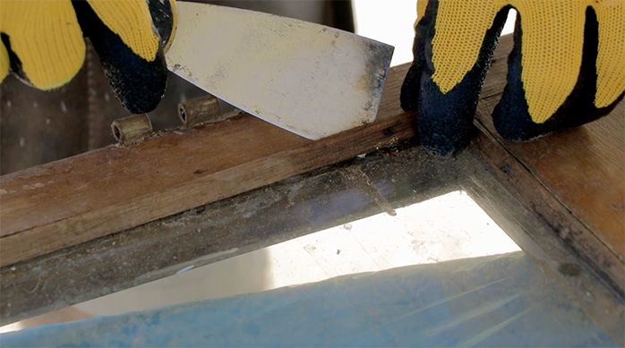 una persona con guantes usa una espátula para sacar los sobrantes de una ventana de madera