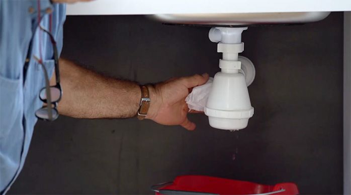 un hombre pasa una toalla absorbente por detrás del sifón de un lavaplatos de cocina para verificar si tiene una filtración