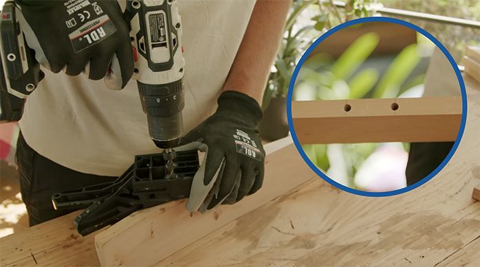 un hombre con guantes hace dos perforaciones con una guía de tarugos en un trozo de madera