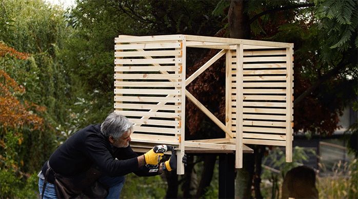 un hombre fija los listones de pino seco cepillado de 1 x 2" en los laterales de la estructura del lugar de almacenamiento para basura en exterior.