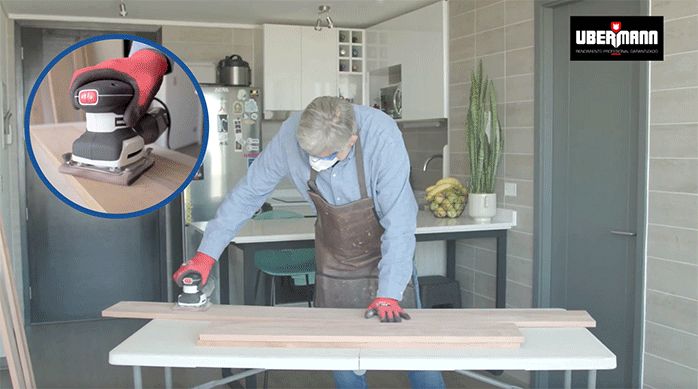 un hombre lija las tablas para hacer el mueble de arrimo con una lijadora roto orbital.