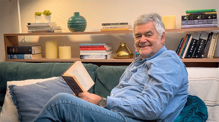 un hombre sentado en un sofá con un mueble de arrimo para libros en la parte posterior, que contiene libros, decoraciones y lámpara.