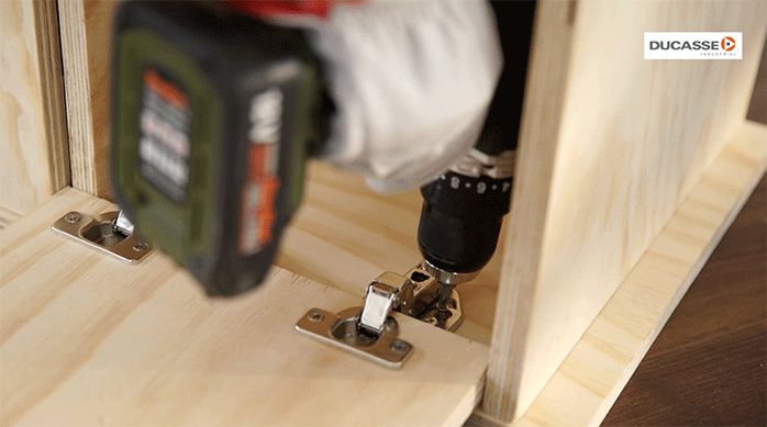 un hombre fija la puerta del mueble atornillando las bisagras al interior de su estructura.