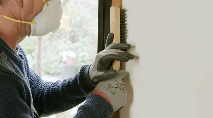 un hombre pasa una escobilla de acero en el marco de una ventana, usando guantes de trabajo y mascarilla.