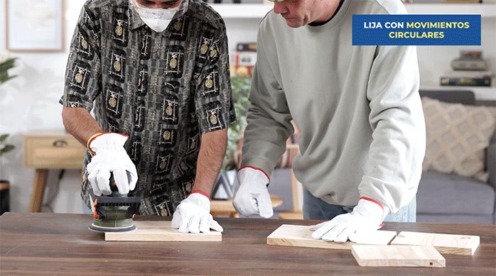 dos hombres con guantes de trabajo. Uno de ellos está lijando un trozo de madera con una lijadora roto orbital.