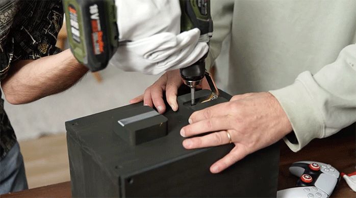 dos personas atornillan 2 ganchos plegables en uno de los laterales del organizador gamer.