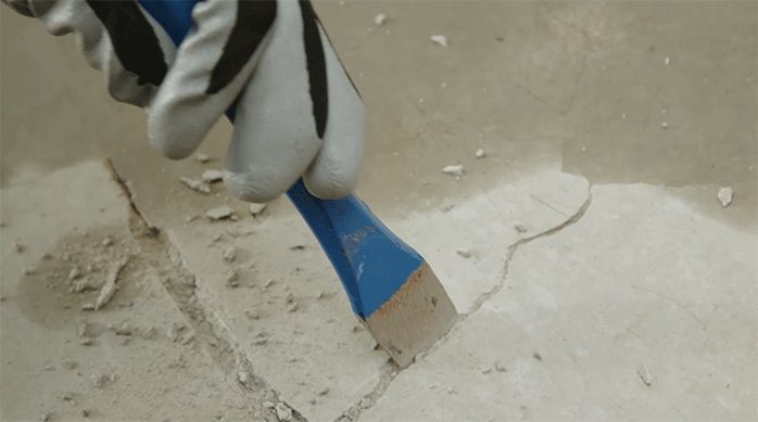 una persona con guantes de trabajo usa un cincel y un martillo para agrandar una fisura en un radier.