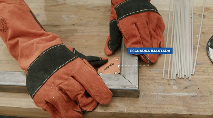 una persona con guantes para soldar fija una unión de perfiles metálicos usando una escuadra metálica.
