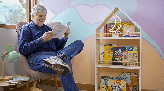 un hombre sentado en un sitial rosado que está puesto en la esquina del rincón de lectura infantil, frente a un librero.