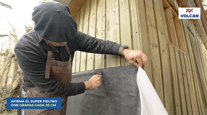 un hombre fija una malla de super fieltro con una engrapadora en la pared exterior de una casa.