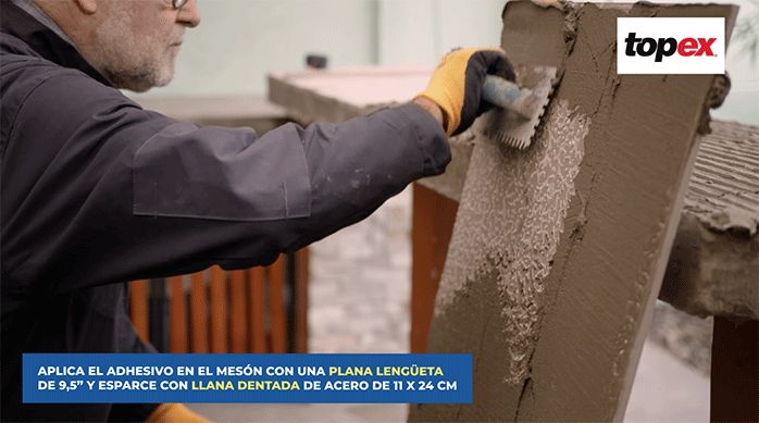 un hombre aplica pegamento para porcelanato en el reverso de una plancha de porcelanato usando una llana dentada.
