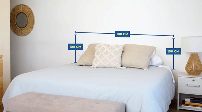 una cama de colores neutros en una habitación de paredes blancas, con una gráfica que muestra las medidas ideales del respaldo de cama.