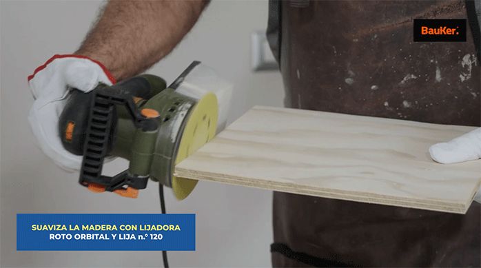 una persona con guantes de seguridad suaviza los cantos de un trozo de madera usando una lijadora roto orbital con lija n.° 120.