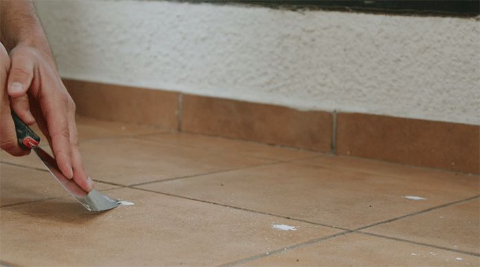 una persona usa una espátula para quitar suciedades pegadas en el piso de cerámica de un balcón.