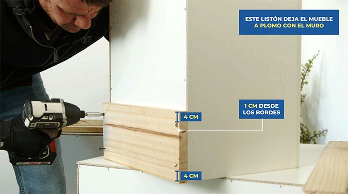 un hombre fija un trozo de madera en la parte posterior de un mueble modular de cocina usando tornillos.
