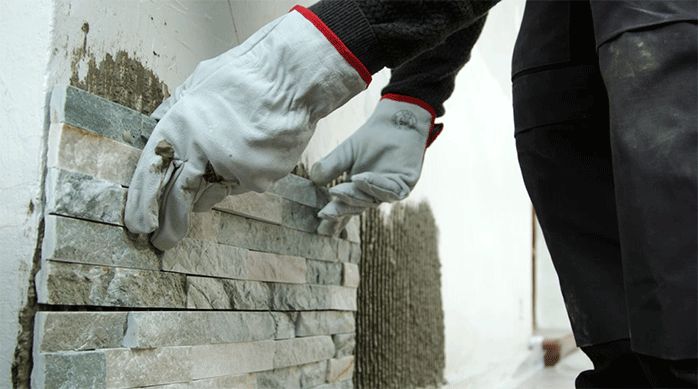 una persona con guantes de trabajo pega una palmeta de revestimiento de piedra natural en el muro de una fachada.