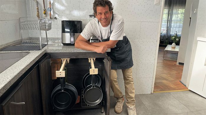 un hombre apoyado en el mesón de una cocina, bajo el cual hay un mueble con las puertas abiertas en que hay correderas en donde cuelgan ollas y sartenes.