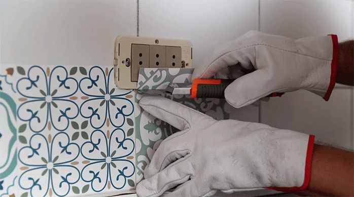 un hombre corta el mosaico adhesivo en el contorno del enchufe, usando un cuchillo cartonero.