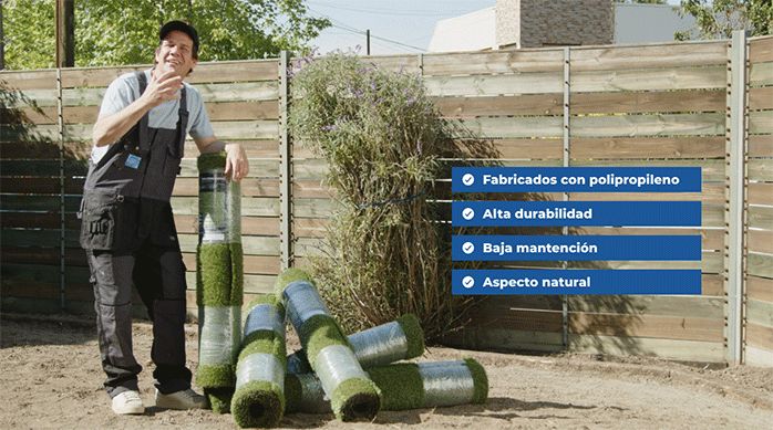 un hombre junto a varios rollos de pasto sintético, en un patio de tierra.
