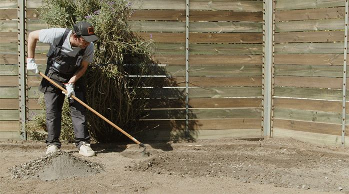 una persona usa un rastrillo para esparcir la mezcla de gravilla y arena de construcción por el piso de tierra.