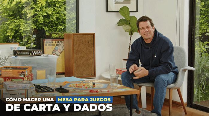 un hombre sentado en una silla, frente a una mesa de centro que tiene juegos de mesa encima, listo para enseñar cómo hacer una mesa para juegos con accesorios.