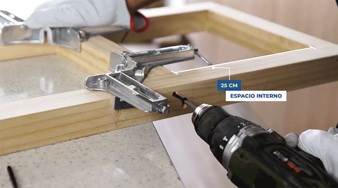 una persona fija el travesaño de un marco de pino seco cepillado usando tornillos y prensa escuadra.