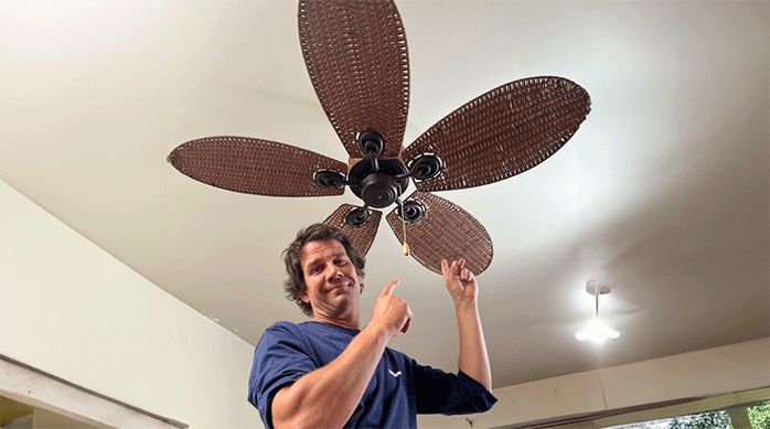 un hombre muestra un ventilador de techo con detalles en ratán recién instalado en un living.