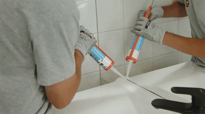una persona aplica un cordón de silicona con la pistola calafatera en la unión entre el lavamanos y el muro del baño.