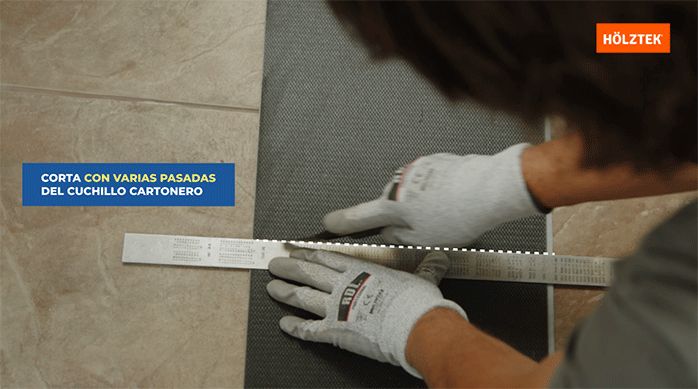 un hombre corta una plancha de piso vinílico por detrás usando un cuchillo cartonero y una regla metálica.