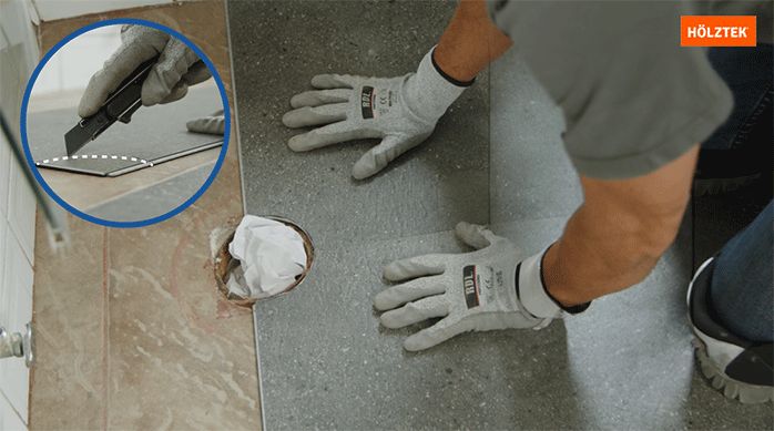 un hombre instala piso vinílico, haciendo los cortes necesarios para dejar espacio al desagüe del baño.