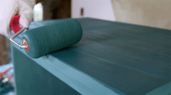 una persona pinta un velador usando un rodillo esponja y pintura verde.