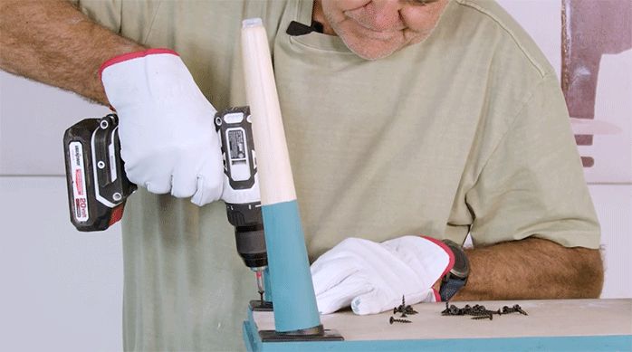 un hombre instala unas patas de madera en la base del velador usando un atornillador eléctrico y tornillos.
