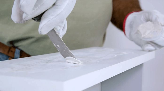 una persona aplica pasta de retape con una espátula en las imperfecciones del cajón del velador.