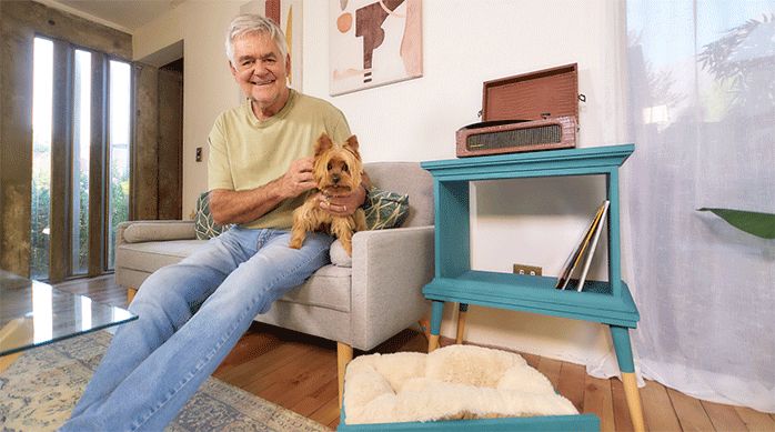 un hombre junto a un perro sentados en un sillón, al lado de un mueble para tocadiscos hecho a partir de un velador antiguo y una cama para mascotas hecha a partir del cajón del velador antiguo.