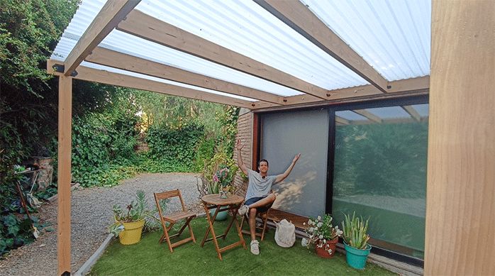 un hombre sentado en un juego de terraza bajo el cobertizo de madera que acaba de construir.