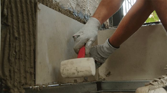 un hombre golpea suavemente una palmeta de porcelanato recién pegada usando un mazo de goma.
