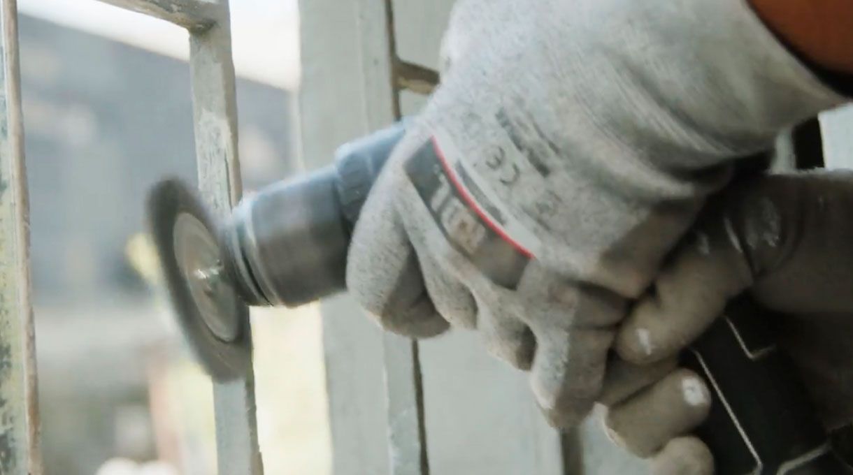 una persona con guantes de trabajo pasa una grata de acero en una reja metálica para eliminar la pintura vieja y el óxido.