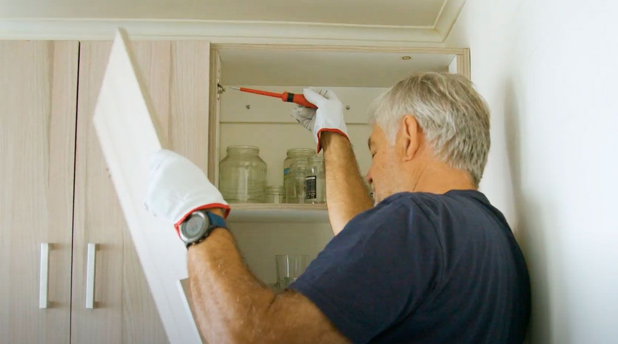 un hombre mayor saca la puerta de un mueble de cocina usando un desatornillador manual.