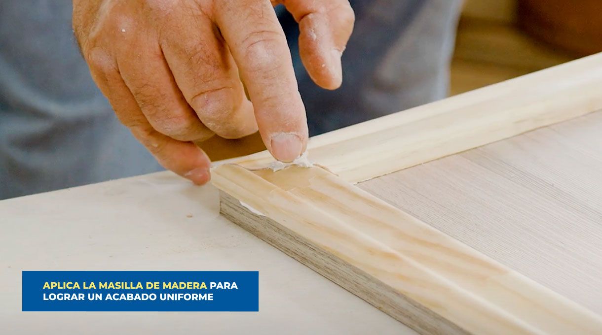 una persona aplica pasta de retape en la unión de 2 trozos de moldura en una puerta de mueble de cocina.