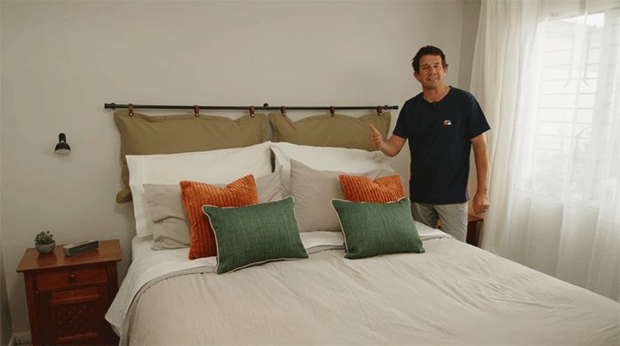 un hombre parado al lado de una cama king con un respaldo hecho de cojines de color beige.