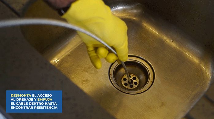 una persona con guantes introduce una sonda destapadora en el desagüe de un lavaplatos.