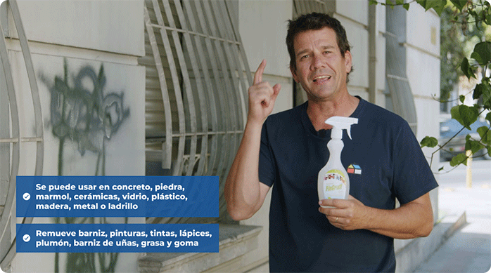 una persona sostiene una botella de removedor de grafitis en la mano y está parado frente a una fachada que tiene rayados.