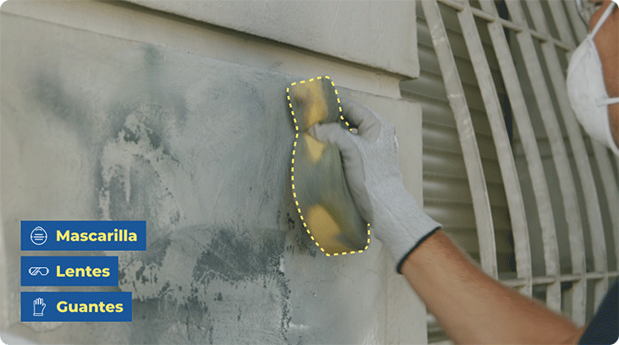 una persona con mascarilla y guantes usa una esponja para eliminar un rayado o grafiti del muro de una fachada.