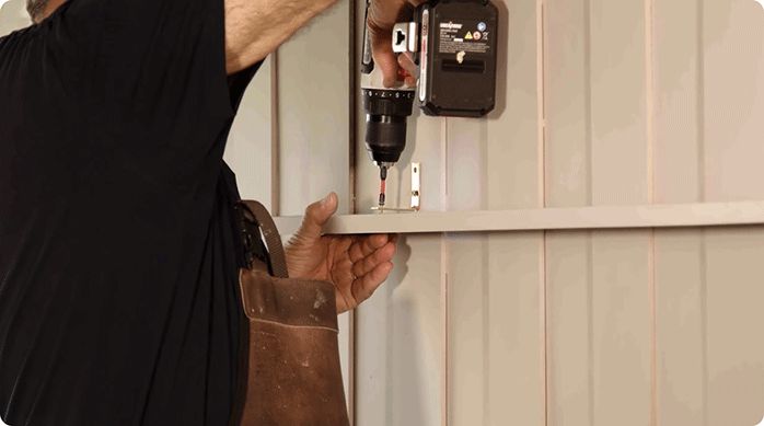 un hombre fija una repisa en una escuadra que está pegada en el muro del dormitorio, usando tornillos.